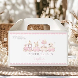 Puede incluir: Caja blanca de golosinas de Pascua con asa, con un tren rosa que transporta personajes de Pascua. La caja está decorada con un borde festoneado rosa y las palabras "Easter Treats Sophie" en una fuente cursiva. También están impresas las palabras "Special Delivery".