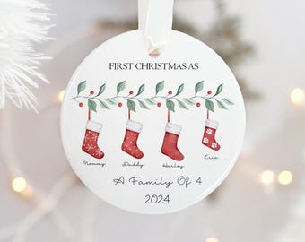 May include: White ceramic Christmas ornament with a green garland and four red stockings. The stockings are labeled "Mommy", "Daddy", "Harley", and "Eric". The ornament also says "First Christmas as a Family of 4 2024".