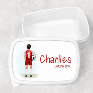 May include: White rectangular lunch box with a lid. The lid features a cartoon illustration of a footballer in a red kit holding a football. The name "Charlies" and "LUNCH BOX" are written in red.