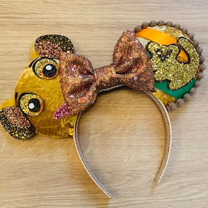 May include: A Simba-themed headband with plush ears and a sequined bow. The ears are designed to resemble Simba's face, with glitter accents. The bow is a shimmering copper colour, and the headband has a gold trim.