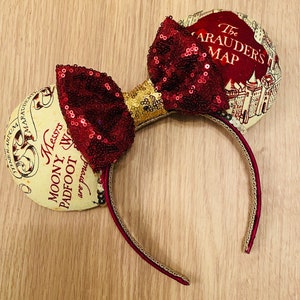 May include: A maroon sequined bow and ears headband with gold accents. The ears are printed with a map design and text that reads "The Marauder's Map" and "Messrs Moony, Wormtail, Padfoot, and Prongs are proud". The headband is maroon with a gold trim.