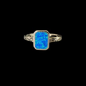 Op de afbeelding: Een zilveren ring met een blauwe opaal-edelsteen gezet in een vierkante bezel. De ringband heeft een decoratief ontwerp.
