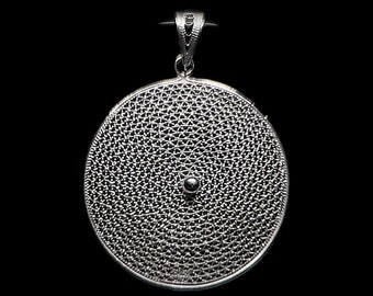 May include: A silver filigree pendant with a circular design. The pendant features a detailed, openwork pattern and a small, centered bead. It has a loop at the top for a chain. The pendant is displayed against a black background.