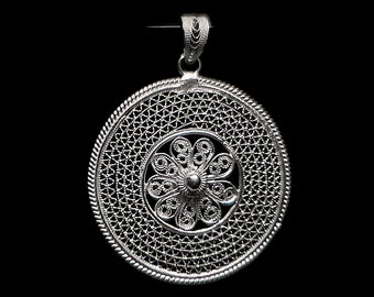 May include: A silver filigree pendant with a circular design. The pendant features an intricate openwork pattern with a central floral motif and a decorative bail for hanging. The pendant is made of a shiny, metallic material and is set against a black background.