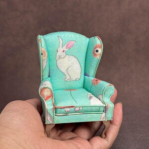 May include: A miniature wingback chair upholstered in a light turquoise fabric featuring a repeating pattern of white rabbits and orange flowers. The small furniture piece has curved armrests and four short, light brown wooden legs.