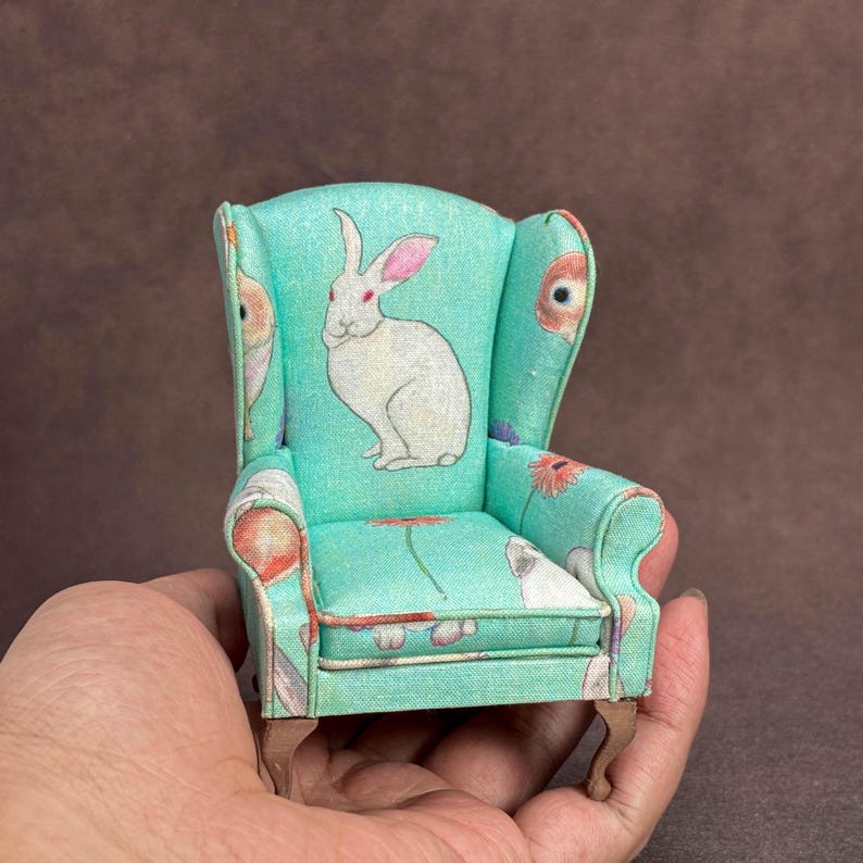 May include: A miniature wingback chair upholstered in a light turquoise fabric featuring a repeating pattern of white rabbits and orange flowers. The small furniture piece has curved armrests and four short, light brown wooden legs.