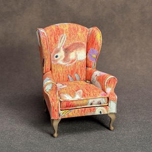 May include: A miniature wingback chair upholstered in a vibrant orange fabric featuring a repeating pattern of rabbits and small purple flowers. The piece has a high back, curved armrests, and four light-colored wooden legs with a cabriole shape.
