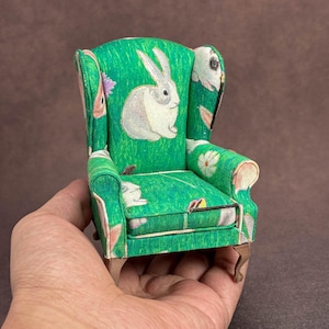 May include: A miniature wingback chair upholstered in green fabric featuring a repeating pattern of white rabbits and small flowers. The chair has a high back, curved armrests, a removable seat cushion, and four small, light brown wooden legs.