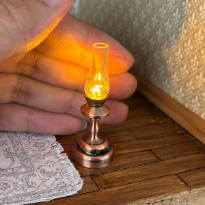 May include: Miniature copper-coloured oil lamp with a clear glass chimney, emitting a warm, yellow glow. The lamp has a detailed base and stem, suggesting a vintage aesthetic. The lamp is a small decorative item.