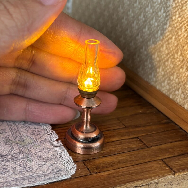 May include: Miniature copper-coloured oil lamp with a clear glass chimney, emitting a warm, yellow glow. The lamp has a detailed base and stem, suggesting a vintage aesthetic. The lamp is a small decorative item.