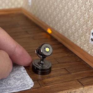 May include: A miniature, dark gray spotlight with a bright white LED. The spotlight is mounted on a small, round base and has a curved arm. The light is positioned on a wooden surface, suggesting a dollhouse or model setting.