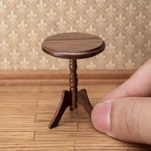 Op de afbeelding: Een kleine, bruine houten tafel met een rond blad en drie poten. De tafel heeft een gedetailleerde, gesneden centrale steun. De tafel wordt door een persoon vastgehouden.