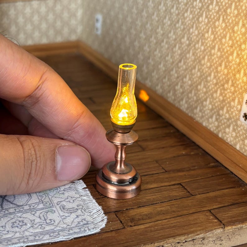 May include: Miniature copper-coloured oil lamp with a clear glass chimney and a warm yellow light. The lamp has a detailed base and stem, and is designed for a dollhouse or miniature setting.