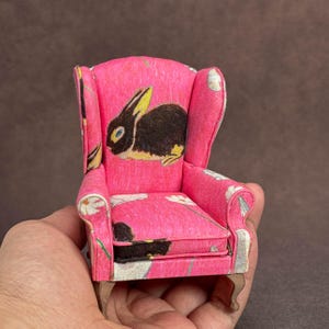 May include: A miniature wingback chair upholstered in bright pink fabric featuring a repeating pattern of black rabbits and white daisies. The small furniture piece has curved armrests, a high backrest, and light-colored wooden legs.
