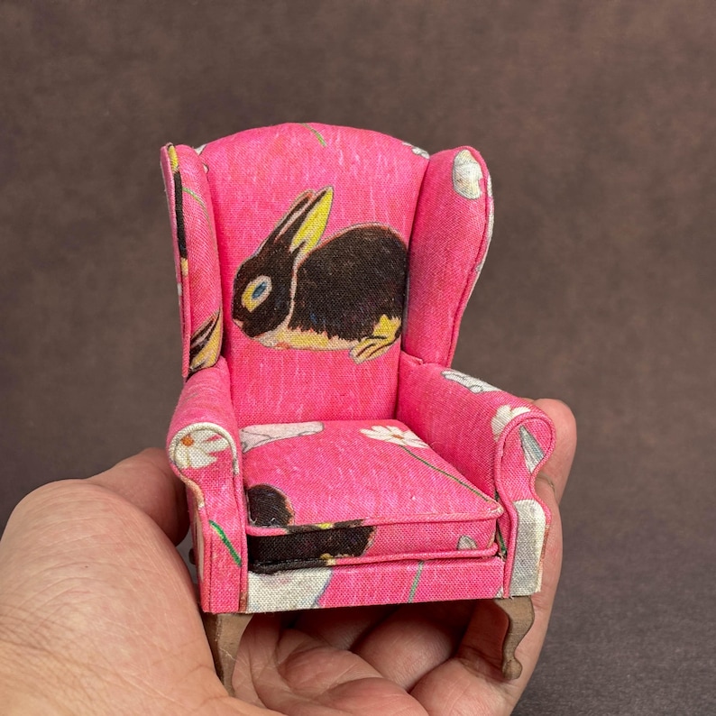 May include: A miniature wingback chair upholstered in bright pink fabric featuring a repeating pattern of black rabbits and white daisies. The small furniture piece has curved armrests, a high backrest, and light-colored wooden legs.