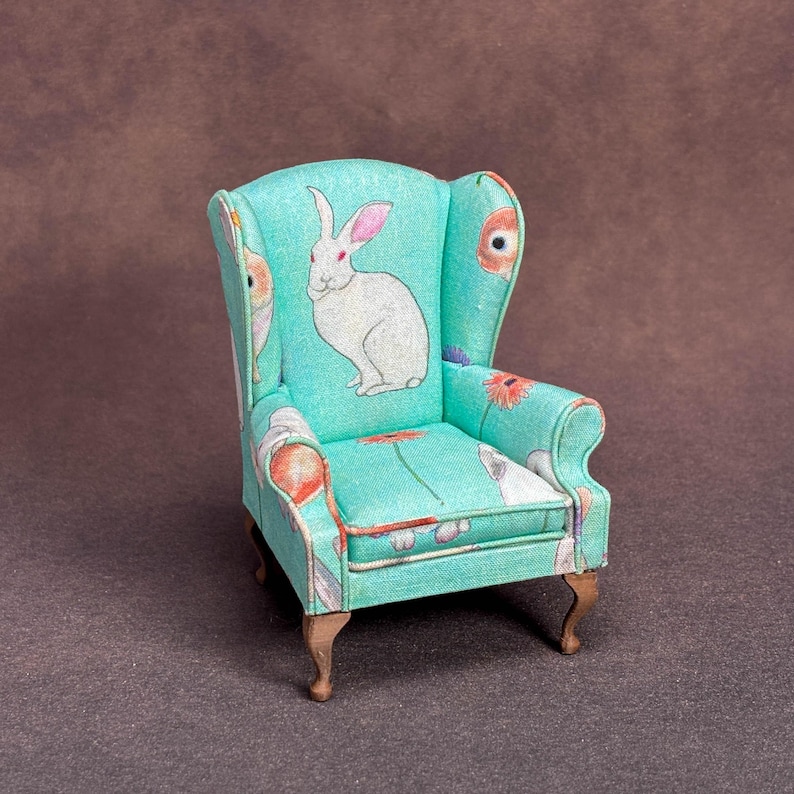 May include: A miniature wingback chair upholstered in a light turquoise fabric featuring a repeating pattern of white rabbits and orange flowers. The small armchair has curved wooden legs and a high backrest with integrated side wings.
