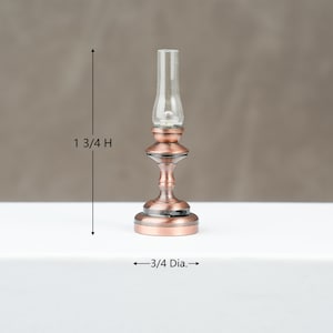 May include: A miniature copper-coloured oil lamp with a clear glass chimney. The lamp is 4.4 cm high and 1.9 cm in diameter. The lamp has a decorative base and a detailed design, suitable for dollhouses or miniature displays.