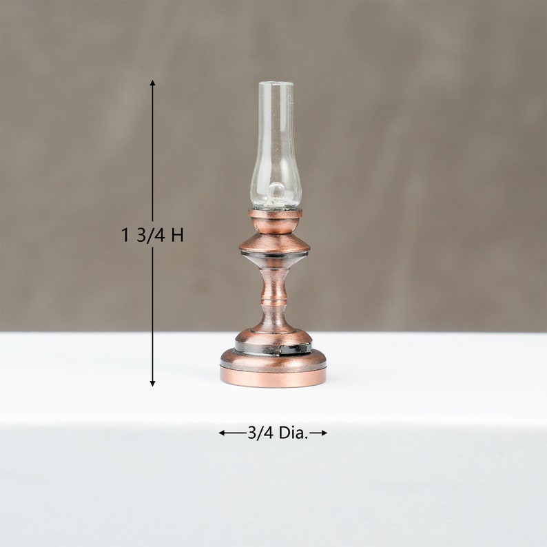 May include: A miniature copper-coloured oil lamp with a clear glass chimney. The lamp is 4.4 cm high and 1.9 cm in diameter. The lamp has a decorative base and a detailed design, suitable for dollhouses or miniature displays.