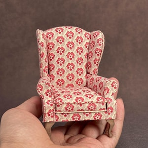 May include: A miniature wingback chair featuring a cream-coloured fabric upholstery with a repeating red floral pattern. The small furniture piece has a high back, curved side wings, padded armrests, and four short, light brown wooden legs.