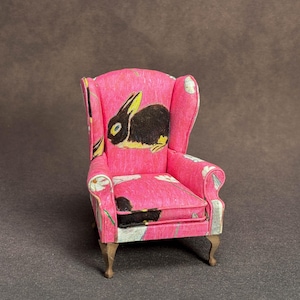 May include: A miniature wingback chair upholstered in bright pink fabric featuring a repeating pattern of black rabbits and white daisies. The small furniture piece has a high back, curved armrests, and four light-colored wooden legs.