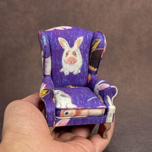 May include: A miniature wingback chair upholstered in a vibrant purple fabric featuring a repeating pattern of white and brown rabbits. The small furniture piece has curved armrests, a cushioned seat, and light brown wooden legs.