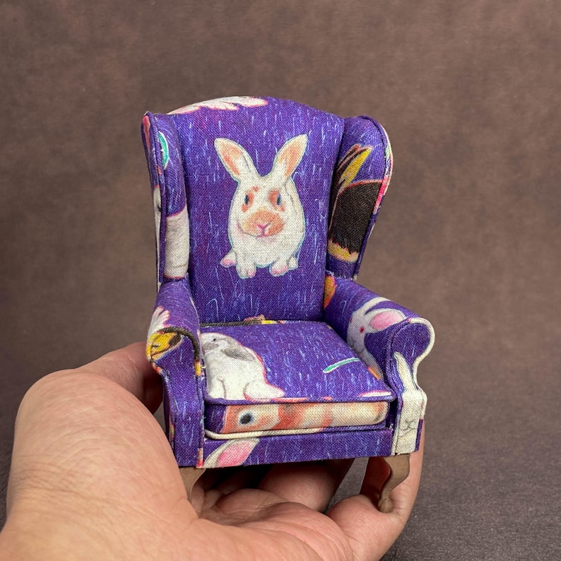 May include: A miniature wingback chair upholstered in a vibrant purple fabric featuring a repeating pattern of white and brown rabbits. The small furniture piece has curved armrests, a cushioned seat, and light brown wooden legs.