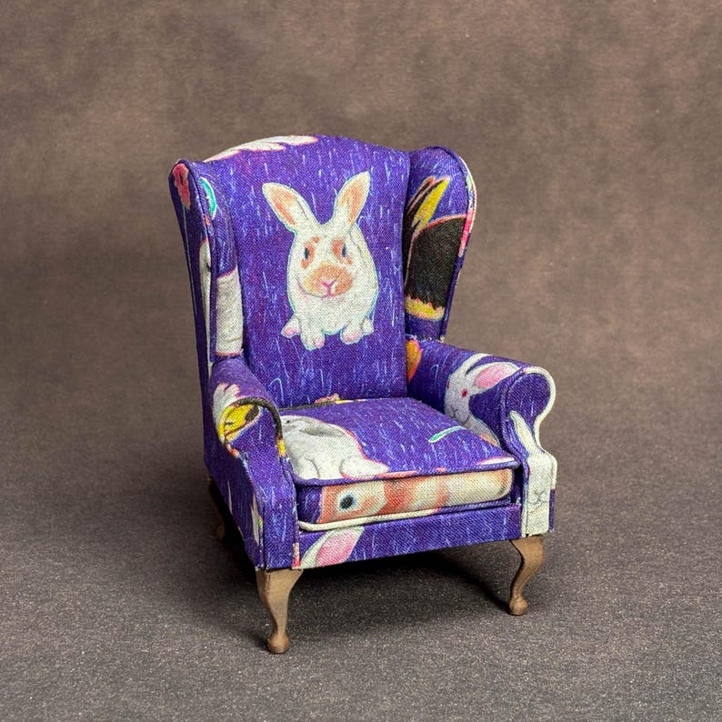 May include: A miniature wingback chair upholstered in a vibrant purple fabric featuring a repeating pattern of white and tan rabbits. The small furniture piece has curved wooden legs and a structured backrest with side wings and padded armrests.
