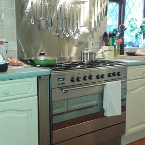 May include: Stainless steel kitchen range hood with lights and a stainless steel backsplash. The backsplash has a rack for hanging kitchen utensils. The range hood is above a stainless steel range with a black top and a white towel hanging on the oven door handle.