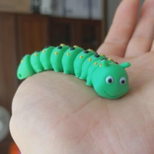 Può includere: Una statuetta a forma di bruco verde brillante con un volto sorridente, occhi mobili e piccole antenne. Il corpo segmentato presenta dettagli neri e gialli. Il giocattolo è tenuto in mano.