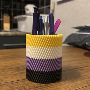 May include: A cylindrical pen holder featuring a ribbed, diagonal texture with horizontal stripes in yellow, white, purple, and black. The container is filled with various pens and writing instruments in shades of blue, silver, purple, and pink.