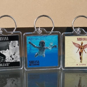 May include: Three clear acrylic keyrings featuring Nirvana album art. The keyrings display album covers for "Bleach", "Nevermind", and "In Utero". Each keyring has a silver-coloured ring for attaching to keys or bags.