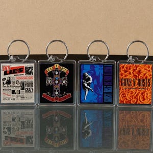 May include: A collection of Guns N' Roses keychains. Each keychain features a different album cover design, including the iconic cross logo and the spaghetti-themed cover. The keychains have silver-colored rings and clear plastic cases.
