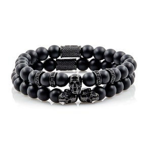 Könnte beinhalten: Zwei schwarze Perlenarmbänder mit Totenkopf-Akzenten. Die Armbänder bestehen aus schwarzen Onyxperlen und haben schwarze Totenkopf-Anhänger aus Metall.