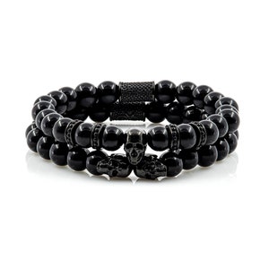 Könnte beinhalten: Zwei schwarze Perlenarmbänder mit Totenkopf-Akzenten. Die Armbänder bestehen aus schwarzen Onyxperlen und haben schwarze Totenkopf-Anhänger aus Metall.