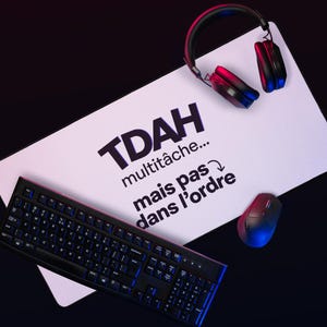 Może przedstawiać: Czarna klawiatura, mysz i słuchawki spoczywają na białej prostokątnej powierzchni z czarnym napisem "TDAH multitâche... mais pas dans l'ordre". Tło jest ciemne z czerwonym i niebieskim oświetleniem.
