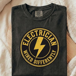May include: Dark gray t-shirt with a round yellow and black graphic that reads "Electrician Wired Differently" with a yellow lightning bolt in the center. The shirt is folded on a wooden surface.