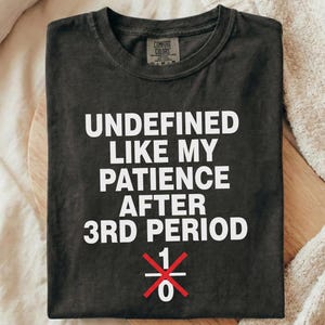 May include: Dark gray t-shirt with the text "UNDEFINED LIKE MY PATIENCE AFTER 3RD PERIOD" in white. Below the text is a red and white graphic. The shirt is folded on a wooden board.