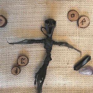 May include: A dark, twisted doll with outstretched arms, crafted from a black, fibrous material. Wooden rune stones and crystals accompany the doll on a textured, tan surface. A piece of wood with a textured surface is in the background.