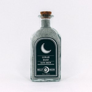 May include: A glass bottle filled with grey bath salts. The label reads "Lunar Dust Bath Brew" with a crescent moon graphic and the text "Wolf Moon 6oz | 180ml".