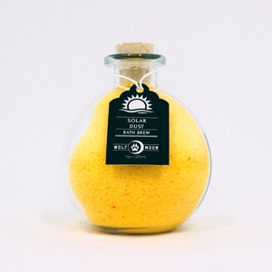Pode incluir: Uma garrafa de vidro cheia de sais de banho amarelos, com uma etiqueta preta que diz "Solar Dust Bath Brew Wolf Moon 132ml".
