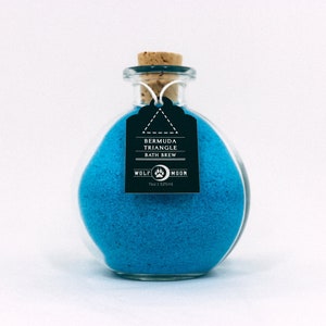 Puede incluir: Una botella de vidrio llena de sales de baño azules. La botella tiene un tapón de corcho y una etiqueta negra que dice "Bermuda Triangle Bath Brew" y "Wolf Moon" con un logotipo de huella de pata.