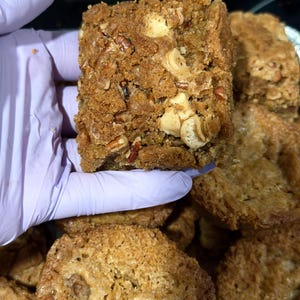Op de afbeelding: Een vierkant gebakje, vastgehouden door een gehandschoende hand. Het dessert is lichtbruin, met zichtbare noten en een kruimelige textuur. Andere soortgelijke bakwaren zijn zichtbaar op de achtergrond.