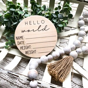 May include: A round wooden sign with the text "HELLO world" in cursive script. The sign has blank lines for "NAME", "DATE", "HEIGHT", and "WEIGHT". The sign is surrounded by white beads and a brown tassel.