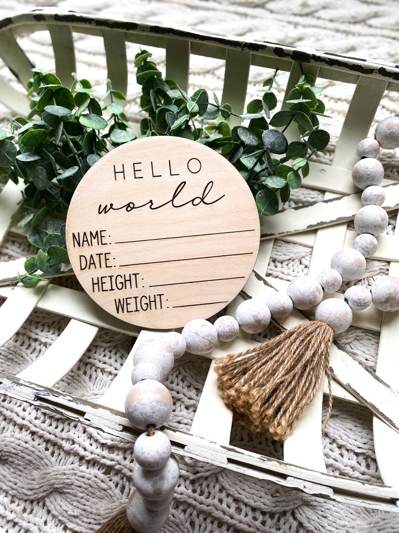 May include: A round wooden sign with the text "HELLO world" in cursive script. The sign has blank lines for "NAME", "DATE", "HEIGHT", and "WEIGHT". The sign is surrounded by white beads and a brown tassel.