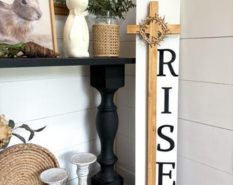 May include: A white wooden sign with the words "He is Risen" in black lettering. A wooden cross with a brown woven crown of thorns is attached to the sign. The sign is leaning against a white wall.