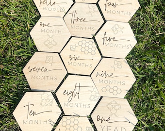 May include: Wooden hexagonal baby milestone cards with a bee theme. Each card features a different month from one to twelve, with the last card saying "one year". The cards are arranged in a circle on a green grassy surface.