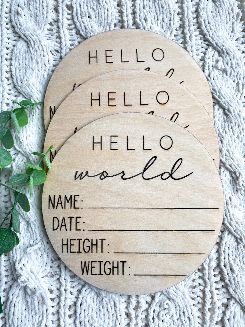 May include: A set of three round wooden signs with the text "Hello World" and blank lines for name, date, height, and weight. The signs are stacked on top of each other.