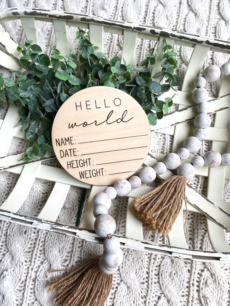 May include: A wooden round sign with the text "HELLO world" and blank lines for "NAME", "DATE", "HEIGHT", and "WEIGHT". The sign is in a white basket with a white and brown beaded garland.