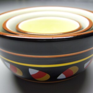 Puede incluir: Un tazón de cerámica negro con un diseño de maíz dulce amarillo, naranja y blanco. El tazón está anidado dentro de otros dos tazones, creando un efecto de capas.
