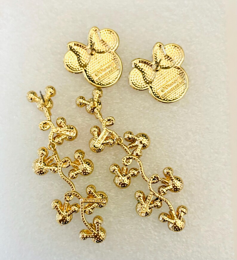 May include: Gold-tone dangle earrings featuring a Mickey Mouse head design. The earrings are made of metal and have a dangling chain of Mickey Mouse heads. The back of the earrings have the text "Disney" and "&copy;Disney" on them.